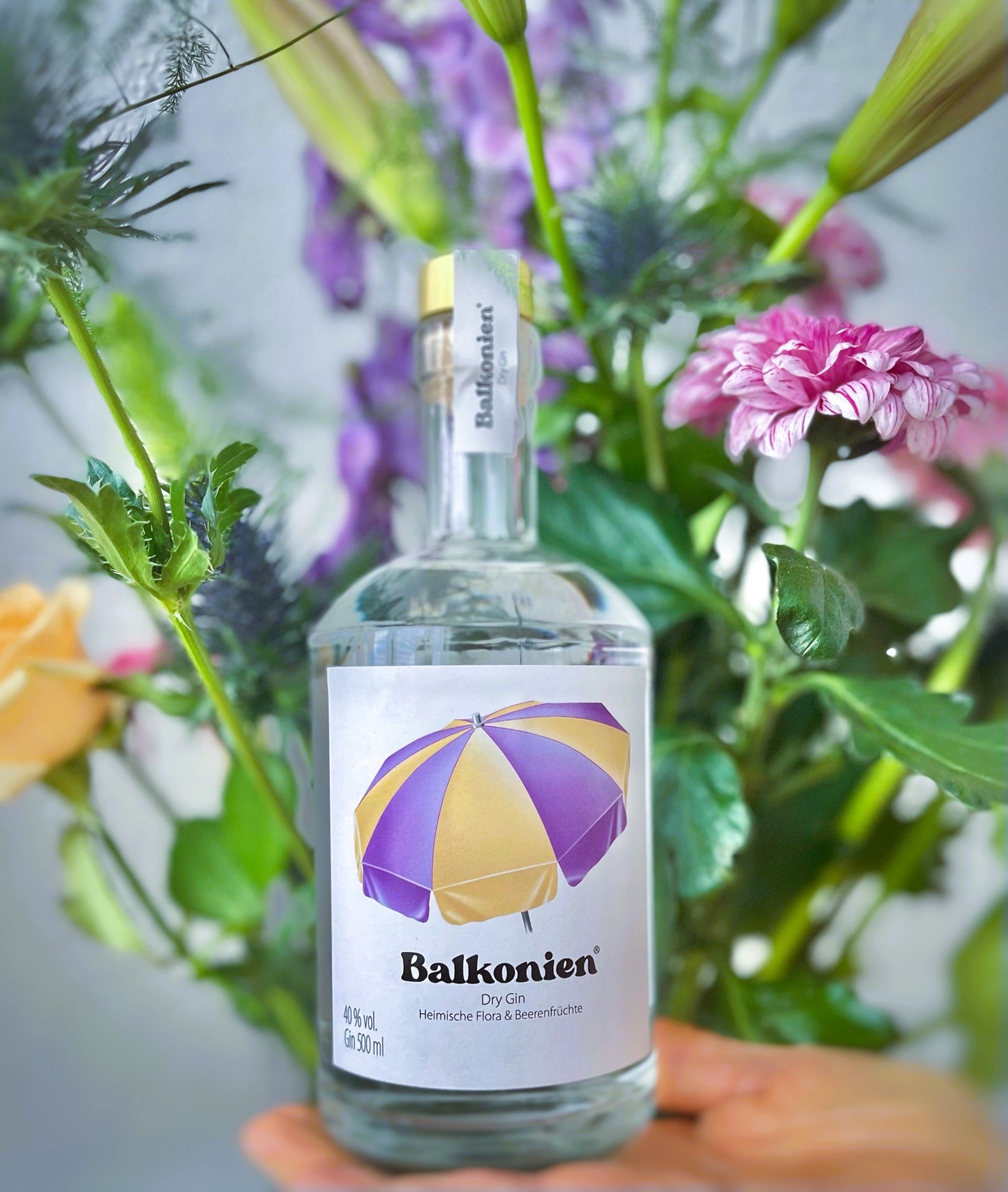 Flasche Balkonien Gin (Heimische Flora & Beerenfrüchte) mit Etikett und lila-gelbem Sonnenschirm, im Hintergrund ein Blumenstrauß.