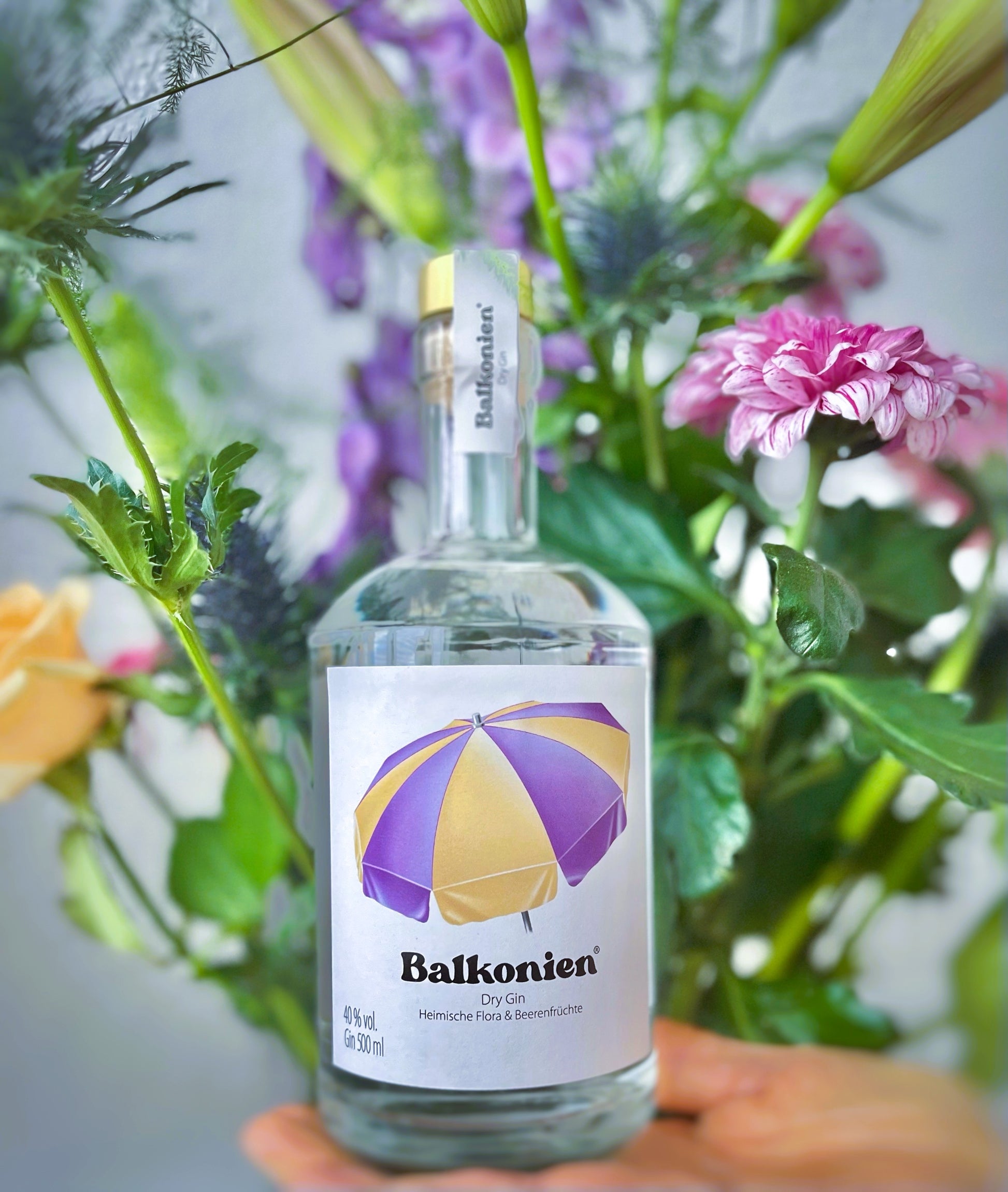 Flasche Balkonien Gin (Heimische Flora & Beerenfrüchte) mit Etikett und lila-gelbem Sonnenschirm, im Hintergrund ein Blumenstrauß.