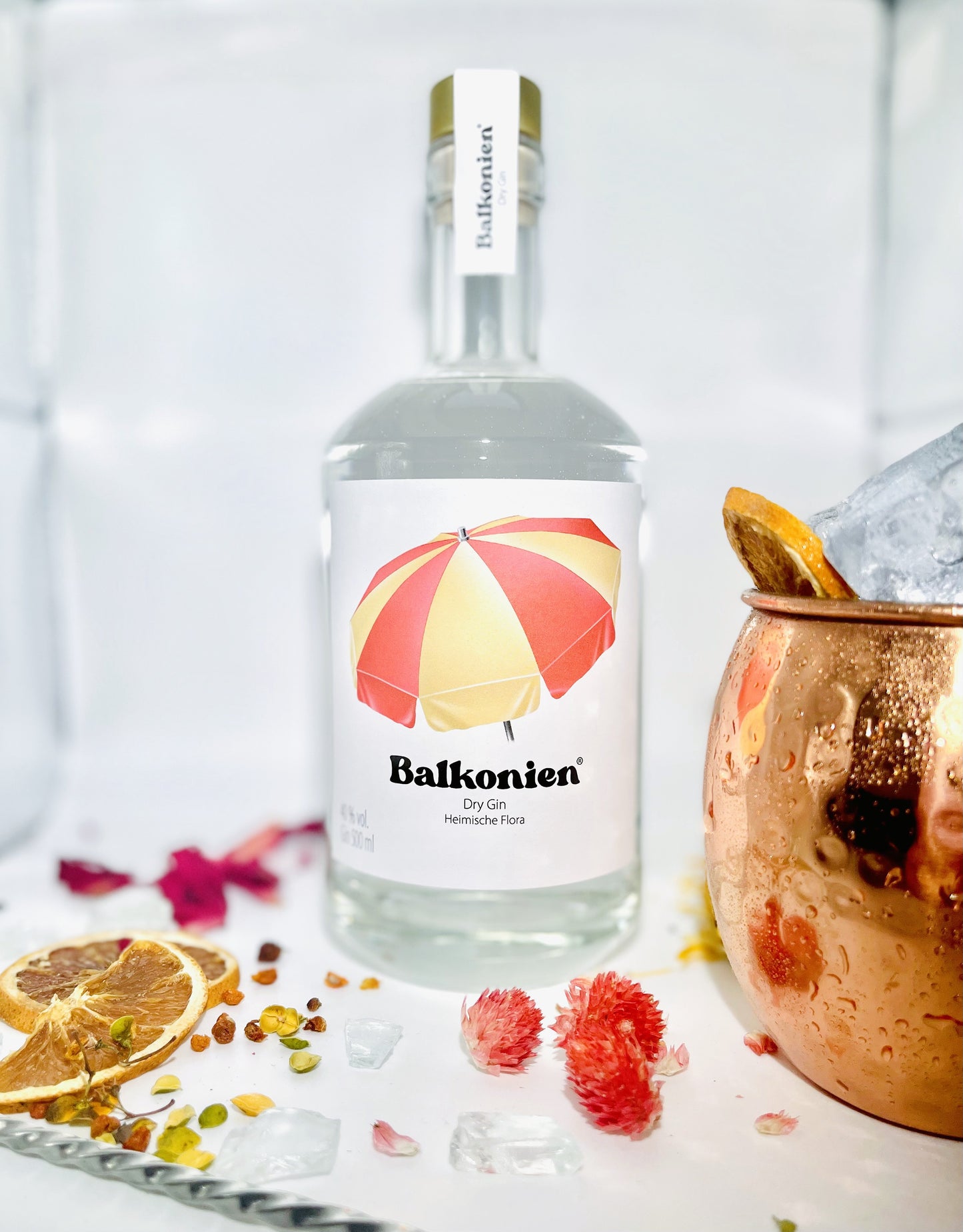 Flasche Balkonien Gin (Heimische Flora) mit Etikett und rot-gelbem Sonnenschirm, umgeben von getrockneten Blüten, Eiswürfeln und einer Kupfertasse mit Eis und getrockneter Orangenscheibe.