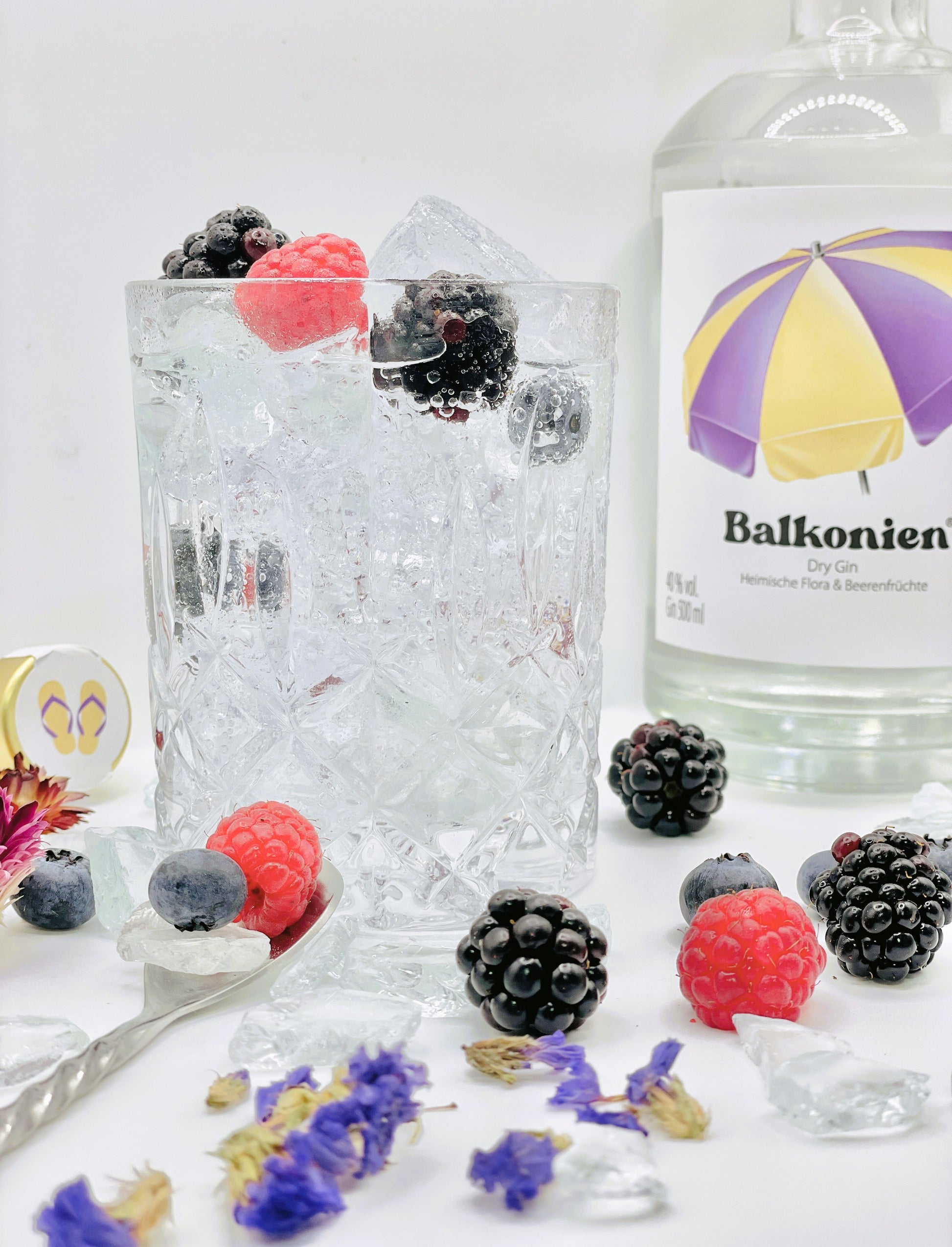 Gin-Getränk in einem Kristallglas mit Eiswürfeln, Himbeeren, Brombeeren und Heidelbeeren in einem Kristallglas, daneben eine Flasche Balkonien Dry Gin sowie verstreute Beeren und Blüten auf weißem Hintergrund.
