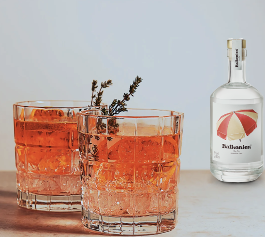 Zwei mit Eiswürfeln und Kräutern garnierte Gläser mit orangefarbenem Gin-Mischgetränk neben einer klaren Balkonien Ginflasche (Heimische Flora) mit rot-gelben Sonnenschirm.