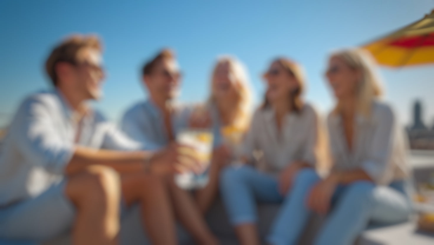 Leicht unscharfes Bild mit mehreren Personen, einige halten Getränke, rechts ein rot-gelb gestreifter Schirm sichtbar.